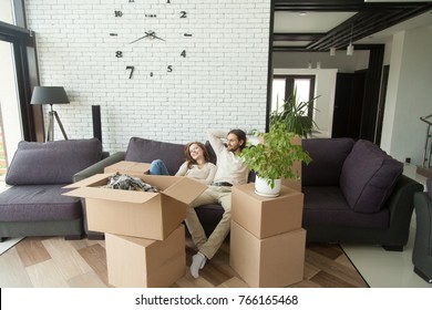 Young Couple Relaxing On Couch Just Moved Into New Home With Cardboard Boxes, Happy Man And Woman Packed Belongings Sitting On Sofa In Living Room, Moving Delivery Service And Relocation Concept