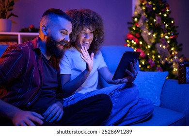 Young couple is relaxing on a sofa in the living room, decorated for the holidays, and having a video call with their family - Powered by Shutterstock