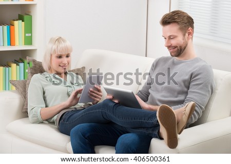 Young Couple Relaxing On Sofa Using Digital Tablets At Home