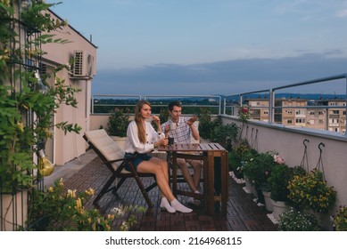 Young couple relaxing outdoors on urban rooftop garden with blooming flowers. Man and woman in casual clothes enjoying sunset and drinking tea or coffee at apartment balcony terrace with city view. - Powered by Shutterstock