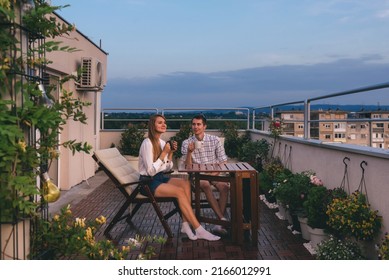 Young couple relaxing outdoors on urban rooftop garden with blooming flowers. Man and woman in casual clothes enjoying sunset and drinking tea or coffee at apartment balcony terrace with city view.
