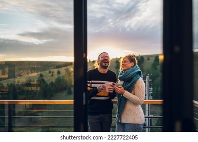 Young couple relaxing outdoors on a balcony. Man and woman in warm clothes enjoying sunrise and drinking tea or coffee at terrace with beautiful nature view. Couple enjoying morning together. - Powered by Shutterstock