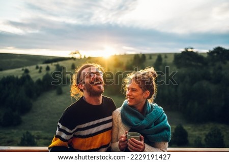 Young couple relaxing outdoors on a balcony. Copy space. Man and woman in warm clothes enjoying sunrise and drinking tea or coffee at apartment terrace with beautiful nature view.
