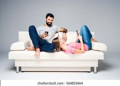 Young Couple Resting On White Couch And Using Smartphones Isolated On Grey