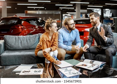 Young Couple With Sales Manager On The Comfortable Couch At The Car Dealership. Young Family Buying Electric Car Dealing Some Formal Things With Salesman In The Showroom