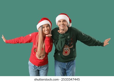 Young couple in Santa hats and Christmas sweaters blowing kisses on green background - Powered by Shutterstock