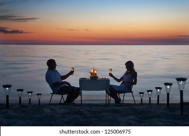 A Young Couple Share A Romantic Dinner With Candles And Wine Glasses On The Sea Sand Beach