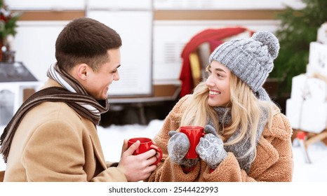 A young couple shares joyful moments while sipping hot beverages in a festive winter setting, surrounded by snow and holiday decorations, showcasing warmth and happiness in the cold. - Powered by Shutterstock