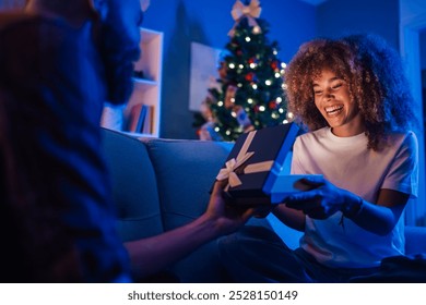 Young couple sharing joy and laughter while opening a christmas present on a cozy blue couch in a festive living room filled with love and christmas lights. Merry christmas and happy new year. - Powered by Shutterstock