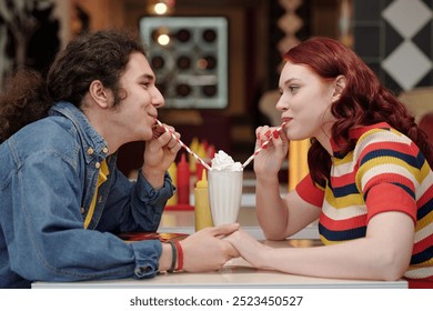 Young couple sharing milkshake in retro-themed diner, both enjoying their time together. Blurred colorful background enhances nostalgic feel, creating warm and cozy atmosphere - Powered by Shutterstock