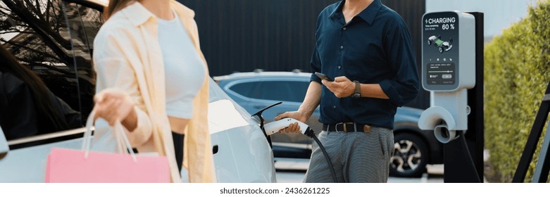 Young couple with shopping bag use smartphone to pay for electricity to recharge EV car battery from charging station at city mall parking lot. Happy couple go shopping by eco car.Panorama Expedient - Powered by Shutterstock