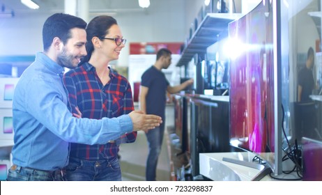 Young Couple Shopping For A New 4K UHD Television Set In The Electronics Store. They're Deciding On The Best Model For Their Happy Family House.