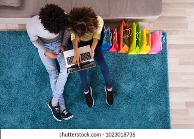 Young Couple Shopping Online On Laptop With Multi Colored Shopping Bags On Sofa