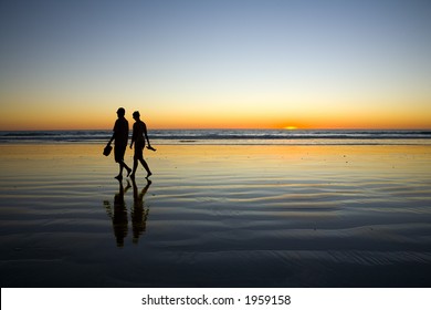 Young Couple In Silhouette Against Sunset Walking At Edge Of Sea On Cable Beach Broome Western Australia. Both Are Barefoot And Carrying Shoes.