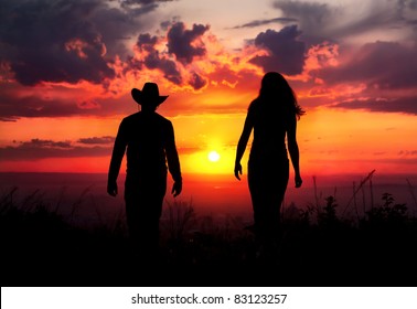 Young Couple Silhouette Walking Outdoors At Sunset Dramatic Sky Background. Man In Cowboy Hat And Woman Nearby