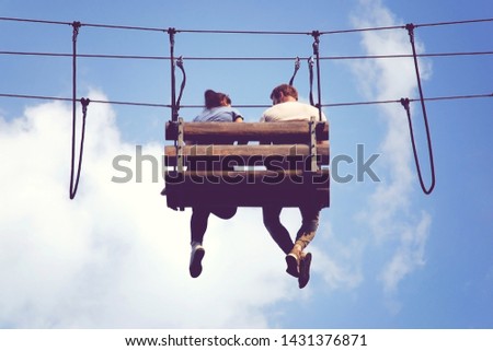 Young couple sit dangling legs on a hanging bench in adventure rope park. Romantic meeting in the skies. 