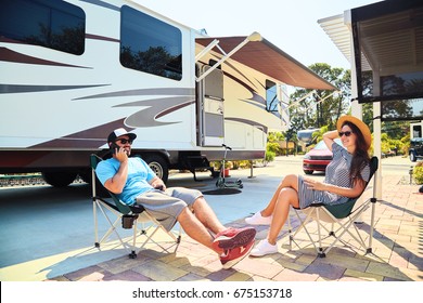 Young Couple Sits Near Camping Trailer,smiling.Men Talks On Mobile Phone And Uses Electronic Device, Woman Relax On Chair Near Car And Palms.Family Spending Time Together On Vacation In Rv Park