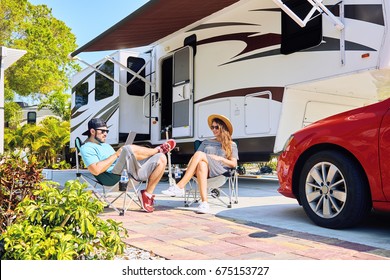 Young Couple Sits Near Camping Trailer,smiling.Men Uses Electronic Device, Woman Relax On Chair Near Red Car And Green Palms.Family Spending Time Together On Vacation In Rv Park