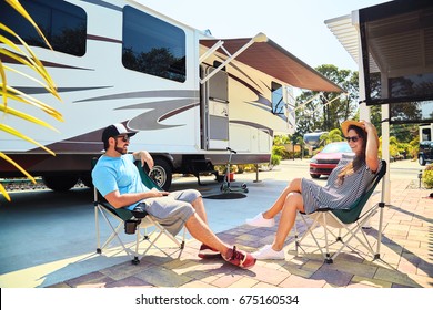 Young Couple Sits Near Camping Trailer,smiling.Men Talks On Mobile Phone And Uses Electronic Device, Woman Relax On Chair Near Car And Palms.Family Spending Time Together On Vacation In Rv Park