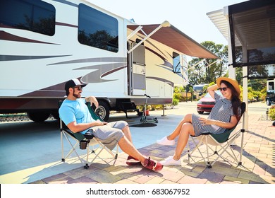 Young Couple Sits Near Camping Trailer,smiling.Men And Woman Talking And Relaxing On Chairs Near Car And Palms.Family Spending Time Together On Vacation In Rv Park