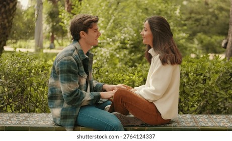 A young couple sits on a patterned park bench, facing each other and holding hands in a warm, sunny outdoor setting. They share a calm, affectionate moment among green trees and shrubs.