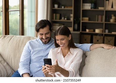 Young Couple Sits On Sofa At Home Smiles Looking At Cellphone Screen, Spouses Having Pleasant Conversation With Family Or Friends Living Abroad Using Video Call Application. Modern Tech, Fun Concept