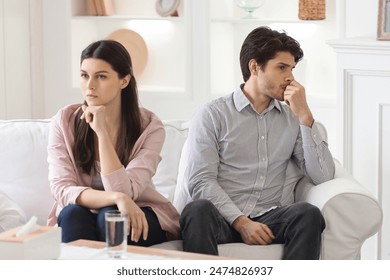 A young couple sits on a sofa in a brightly lit living room. Both appear to be lost in thought, staring off into the distance with serious expressions. Crisis in relationships