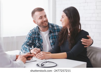 A young couple sits at a table in a doctors office, engaged in conversation with the doctor off-camera. The man is wearing a plaid shirt and the woman is wearing a black long-sleeve shirt. - Powered by Shutterstock