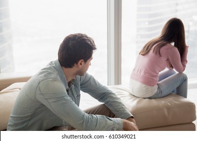 Young couple sitting apart indoors after quarrel. Offended girl ignoring boyfriend, looking away, serious man thinking about problem in relationships, family conflict, misunderstanding between teens 