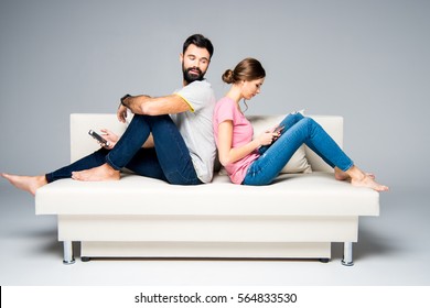 Young Couple Sitting Back To Back On White Couch And Using Smartphones Isolated On Grey