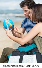 Young Couple Sitting At The Beach Planning Their Holiday Pointing Excitedly At A Globe As They Choose Their Next Destination