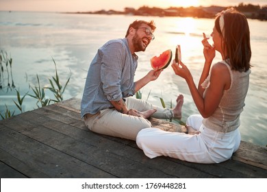 junges Ehepaar, das bei Sonnenuntergang am Fluss sitzt, spricht, lächelt, lacht, Wassermelone isst 