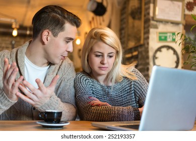 Young Couple Sitting In The Cafe With Laptop Making Some Important Decisions. The Foreground Is Blurred, Narrow Depth Of Field, Natural Light. Soft Focus On The Male Person