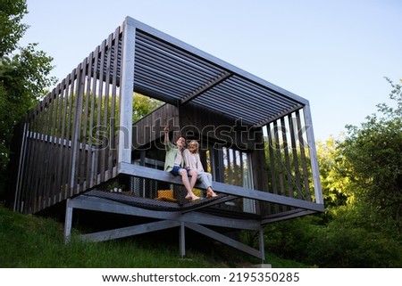 Young couple sitting and cuddling in hammock terrace in their new home in tiny house in woods, sustainable living concept.