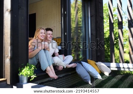 Young couple sitting with cup of coffee and cuddling in terrace in their new home in tiny house in woods, sustainable living concept.