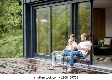 Young couple sitting with cups on the terrace of the modern house enjoying beautiful view outdoors - Shutterstock ID 1108469735