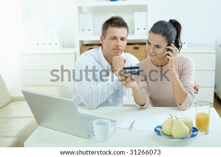 Young couple sitting at desk shopping on internet from home. Calling phone, using laptopcomputer and paying with credit card.