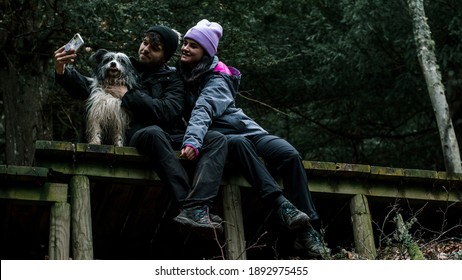 The Young Couple Are Sitting Down And Take Out A Selfie On A Wooden Plank Road In The Forest. Next To Them Is Their Shepherd Dog.