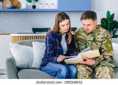 young couple sitting at home in the living room handsome soldier in military camouflage uniform shows his wife photographs of memories of military civil service after returning from the army - Powered by Shutterstock