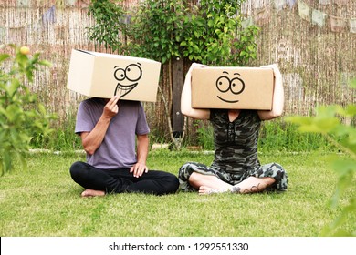 Young Couple Sitting In The Meadow With Boxes Over Their Heads Prank