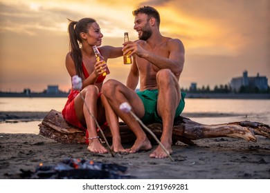 Young Couple Sitting Near Campfire On The Beach At Sunset And Drinking Beer.