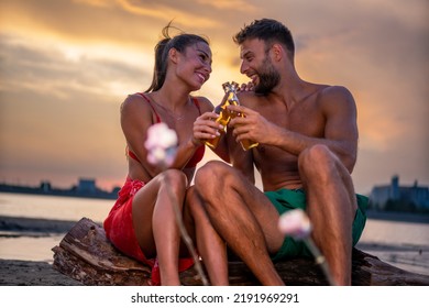 Young Couple Sitting Near Campfire On The Beach At Sunset And Drinking Beer.