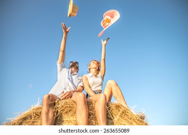 Young Couple Sitting On A Bale Of Hay And Throwing Hats In The Sky