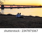 a young couple sitting on the banks of the Mississippi River at Greenbelt park at sunset with yellow winter grass and bare winter trees and a bridge over the water in Memphis Tennessee USA