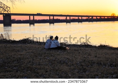 a young couple sitting on the banks of the Mississippi River at Greenbelt park at sunset with yellow winter grass and bare winter trees and a bridge over the water in Memphis Tennessee USA
