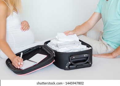 Young couple sitting on bed and packing baby clothes. Pregnant woman hand holding notebook with list and man checking things. Preparing for hospital childbirth or traveling.  - Powered by Shutterstock