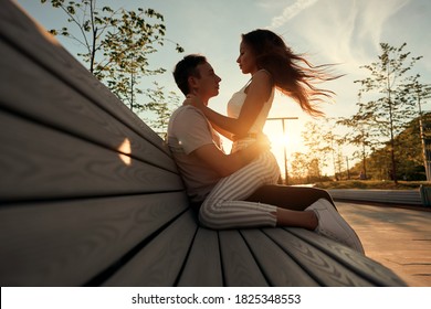 A Young Couple Is Sitting On A Bench At Sunset. The Lady Sits On The Guy's Lap And Looks So Contented And Happy.