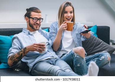 Young Couple Sitting On The Couch, Drinking Coffee And Watching Tv