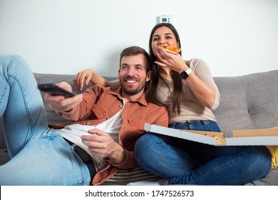 Young couple sitting on a couch at home, eating pizza, watching TV