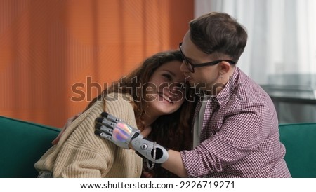 Young couple sitting on couch and hugging. Man with bionic arm. Boyfriend with robotic hand prosthesis embrace smiling girfriend